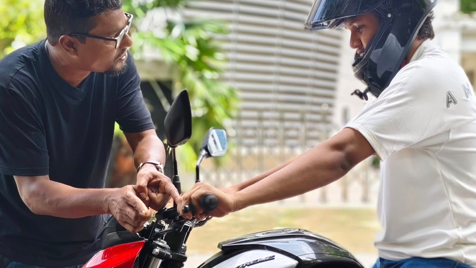 Two men with a motorcycle, one adjusting something on the bike.